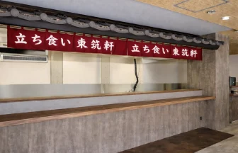 折尾本社を全面リニューアルした東筑軒「ロードサイドモデル1号店」の立ち食いカウンター。伝統のエンジ色の暖簾と瓦屋根のデザインを継承し、旅の情熱を感じさせるモダンな店内。