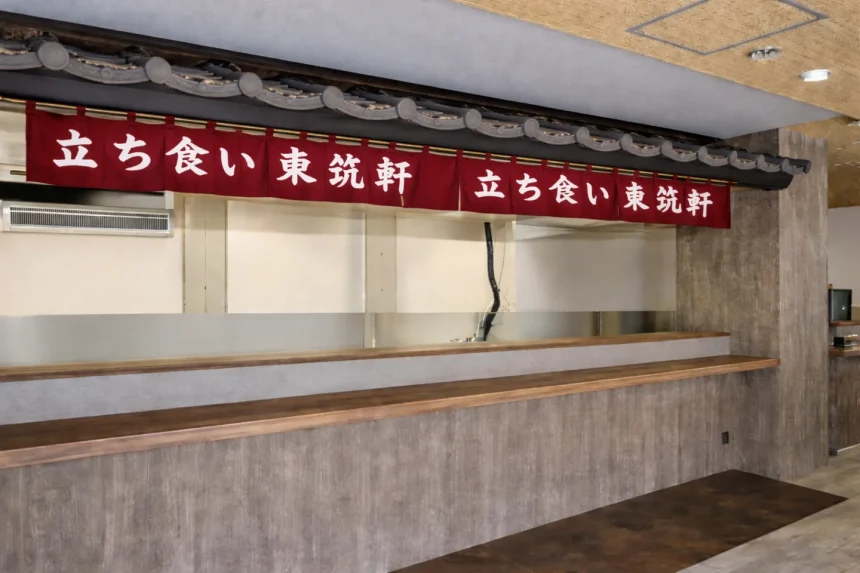 折尾本社を全面リニューアルした東筑軒「ロードサイドモデル1号店」の立ち食いカウンター。伝統のエンジ色の暖簾と瓦屋根のデザインを継承し、旅の情熱を感じさせるモダンな店内。