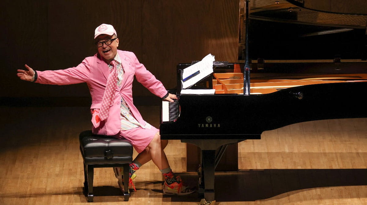 鮮やかなピンク色のスーツとキャップ姿で、笑顔でピアノに向かう大江千里。ポップとジャズを自在に融合させる“三代目”としての自由で楽しげな演奏パフォーマンスの様子。