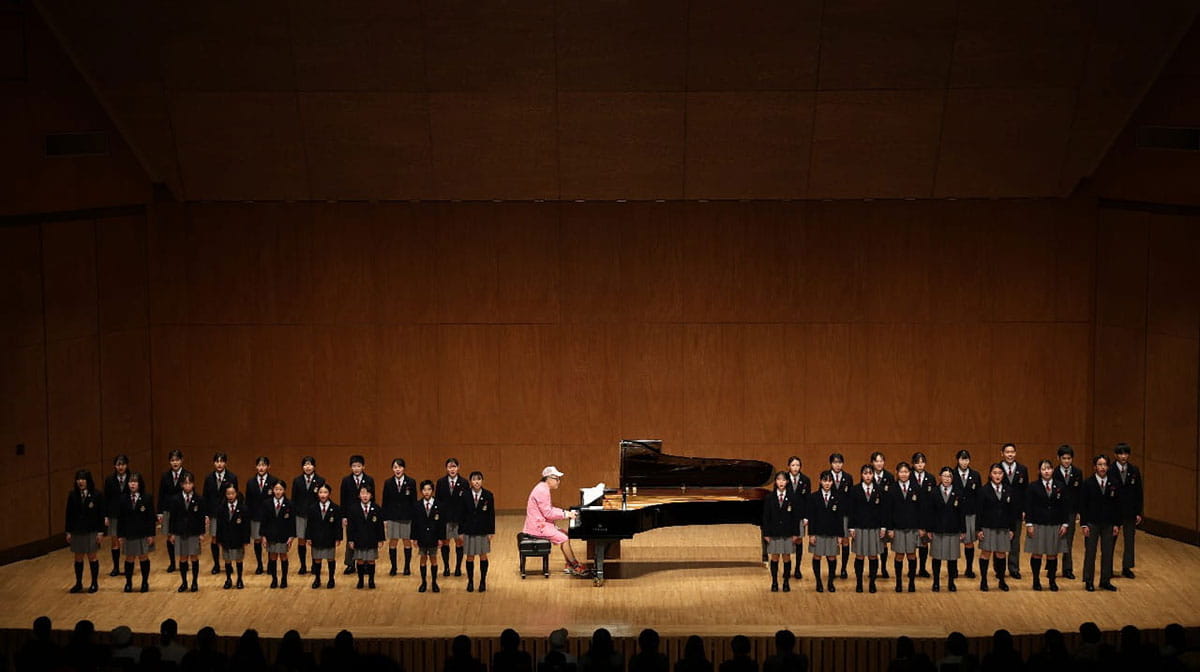 クラシックホールを舞台に、グランドピアノを奏でる大江千里と、その両脇を固める少年少女合唱団(制服着用)の共演風景。世代を超えた音楽の響き合いを感じさせるコンサートの全景。