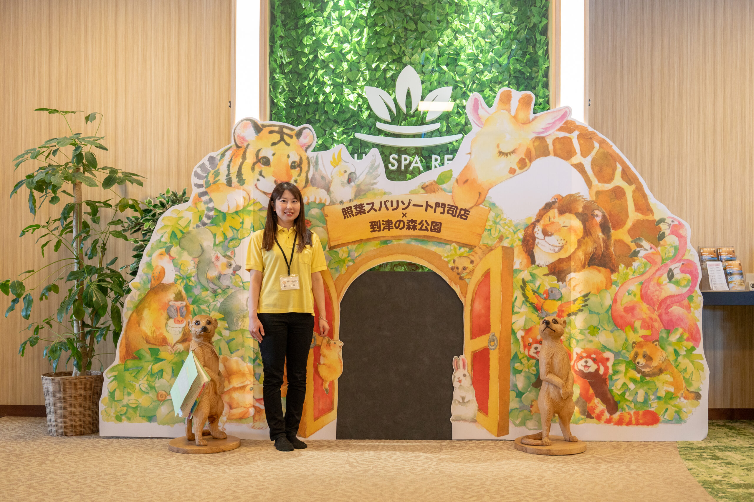 照葉スパリゾート門司店×到津の森公園コラボのフォトスポット前に立つ女性スタッフ。水彩タッチで描かれたキリンやライオン、トラ、レッサーパンダなどの動物イラストに囲まれた大型パネルが設置され、入口には動物園のような扉のデザインがあしらわれている。